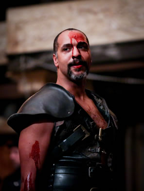 Coriolanus in black armor-like leather vest, blood on his arm, and blood dripping down his face from his forehead.