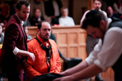 Antonio in orange prison overalls and a halter around his neck with Gratziano in red jacket standing by, hand on Antonio's shoulder. Out of focus at the front is the bowed-headed Shylock packing, and audience members are seen in the background along with Bassanio standing by the wood wall.