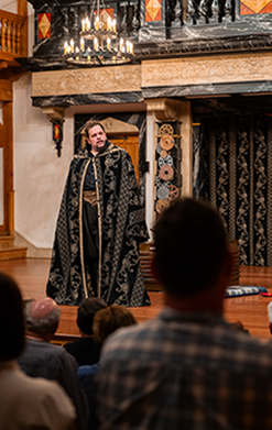 ASC-Twelfth_Night-32-206 In photo taken from within the audience, Blake Henri stands on stage wearing a silver embroadedred black cloak the exact design of the curtain at the bak of the stage behind him. Also visible are the gear-covered pillars, the bottome of the theater's balcon in marbalized wood, and a chandelier.