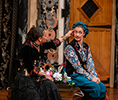 Angelique Archer as Olivia in a long black dress with black jacket embroidered with flowers and Summer England as Viola disguised as Cesario wearing a coloruflly flowered shirt, purple vest, royal blue pants and blu cap sit on each side of a trunk with flowers between them. Olivia is reaching across to stroke Cesario's temple as Viola/Cesario looks uncomfortably to the audience.