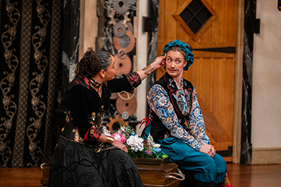 ASC-Twelfth_Night-32-347 Angelique Archer as Olivia in a long black dress with black jacket embroidered with flowers and Summer England as Viola disguised as Cesario wearing a coloruflly flowered shirt, purple vest, royal blue pants and blu cap sit on each side of a trunk with flowers between them. Olivia is reaching across to stroke Cesario's temple as Viola/Cesario looks uncomfortably to the audience.