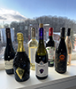Seven bottles of wine in two staggard rows on a window sill with a winter landscape through the window of buildings in front of a hill comprising bare trees. The wines are, from left: Red Earth Matchbook Petit Sirah, Astoria-Galie Prosecco, Domain Gavoty Grand Classique Vermentino, Vignamato "Violetta," Muse "Urania," and Baudry Dutour Chnino Rosé.