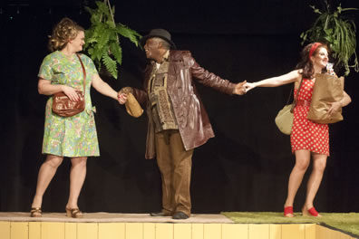 Falstaff in brown leather jacket and brown pants and shirt and hat holding a bagged liquor bottle holds the hands of Mistress Ford in green-patterned  knee-length dress and brown purse, and Mistress page in white polka dot red thigh-length dress, holding a bag of groceries, with hanging planters of ferns behind. 