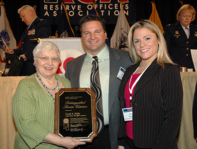 Carol holds the ROA Distinguished Service Citation plaque in her hands as she stands next to Matt, in tie and jacket, and Julie, in dark gray pants suit with red blouse. In the background is the podium for the Reserve Officers Assocation luncheon.