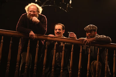 Toby, with elbow on balcony railing, Aguecheek holding a flask and croutched behind the balcony railing, and Fabian with checkered dog cap next to him.