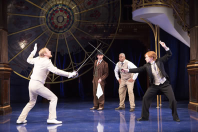 Sir Andrew is in classic white fencing outfit, Viola in a three-piece suit, with short red hair and a fake mustache, Feste holds a handgerchief, and Belch in tan three piece suit without the jacket stands crouched with hands on hip. The huge circular stained glass window is in the backround, and the stairs to the side.