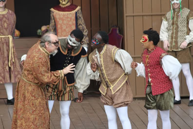 Capulet in long gold and red patterned robe with gold rope belt leans over to talk closely with Benvoloio in black waistcost and black, gold and green striped breeches, Mercutio in gold brocade waistcoat and breaches, and Romeo in red quilt wastecoat and green breeches. All are wearing masks, and all three young men are in white blousy shirts and white tights.