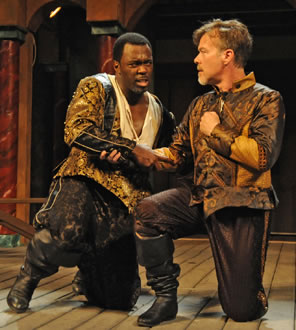 Othello on one knee clasps right arms with Iago, also on one knee and holding a fist to his heart as he swear allegiance,, both in gold-brocaded Renaissance Venitian coats