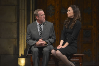 Sitting on a bench with a stone wall and large wooden door in the background, Prime Minister Evans in gray suit and shirt, black and white striped tie, hands folded on a black notebook, has his head turned as he talks to Kate sitting sideways, legs crossed demurely, hands pressing into her lap, a plain blck dress and a far-away look of contemplation.