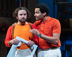 Antipholus in orange polo shirt and white pants has his right hand around the shoulder of dromio wearing multi-orange sweatshirt, suspenders, and white cargo pants and holding the frame of a destroyd gift box top.