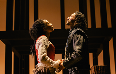 STC_CST-Hamnet-3 With the shadowed black outline of the wood superstructure behind them, Agnes in Simple Elizabethan dress and William in Elizabehtn leather jacket hold hands. He's looking up to the heavens, she's looking up toward his eyes.