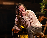 William sits on the center wood structure's lader, his head turned to his left with his chin resting on his left hand and his left elbow resting on his left knee. His in a white Elizabeth shirt. In the background, Agnes is tending to something.