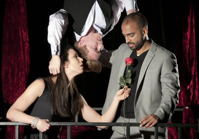A young woman holds a rose up to a man while another man hanging upside down from above has his hands behind the heads of the other two
